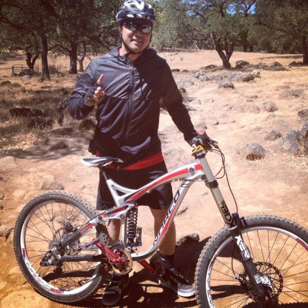 A person wearing a helmet and sunglasses stands next to a mountain bike on a rugged, gravelly trail surrounded by trees. They are smiling and making a hand gesture while dressed in athletic wear, including a black jacket and shorts. The background features a natural landscape with rocky terrain and sparse vegetation. Rockville Park mountain bike trail.