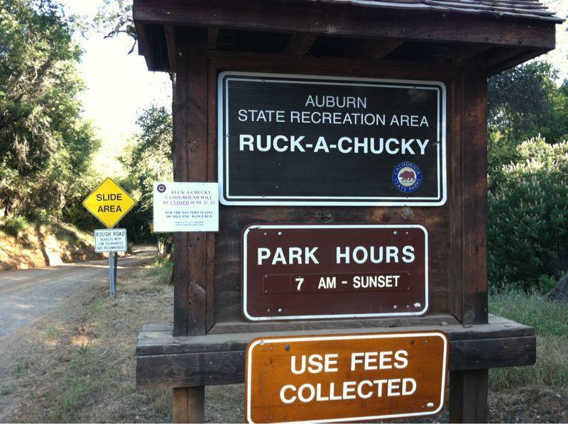 Signage for Auburn State Recreation Area at Ruck-A-Chucky, featuring park hours from 7 AM to sunset, a warning for a slide area, and information about use fees. The sign is situated near a dirt road surrounded by trees. Foresthill Divide mountain bike trail.