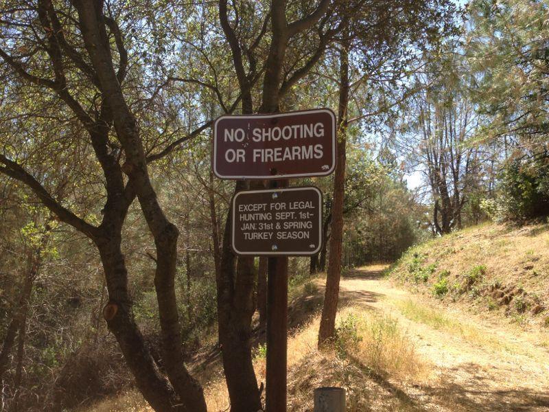 Sign indicating "No Shooting or Firearms" with exceptions for legal hunting seasons, located along a dirt path surrounded by trees. Foresthill Divide mountain bike trail.