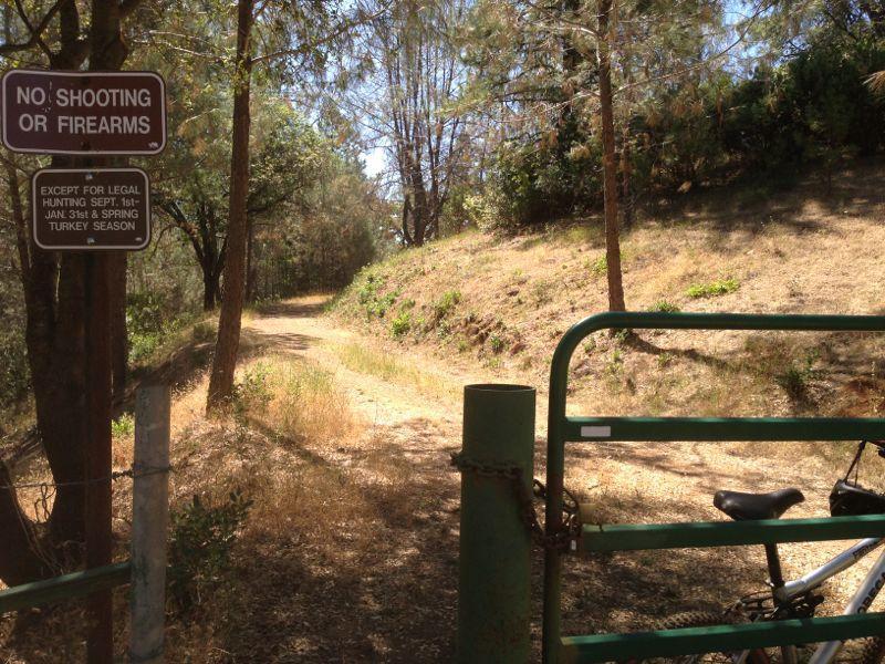 Alt text: A green gate secured with a chain and padlock stands at the entrance of a dirt path surrounded by trees. A sign on the gate reads "No Shooting or Firearms," with additional text regarding legal hunting seasons. Summer vegetation is visible along the trail. Foresthill Divide mountain bike trail.