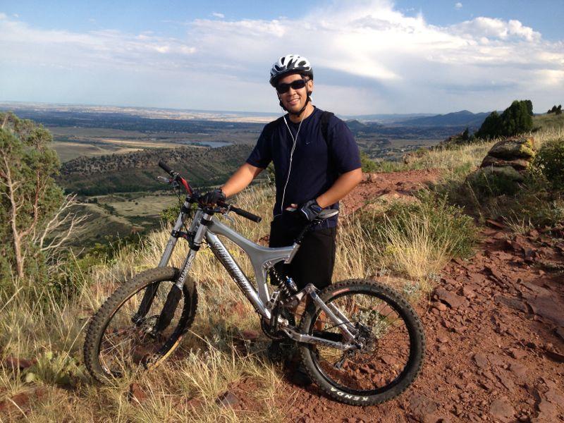 A mountain biker in a black helmet and sunglasses smiles while standing next to a silver mountain bike on a rocky trail. The background features a scenic view of hills and valleys under a partly cloudy sky. The cyclist is dressed in a blue t-shirt and black shorts, with earbuds visible, suggesting a moment of enjoyment during a ride. Red Rocks / Dakota Ridge mountain bike trail.