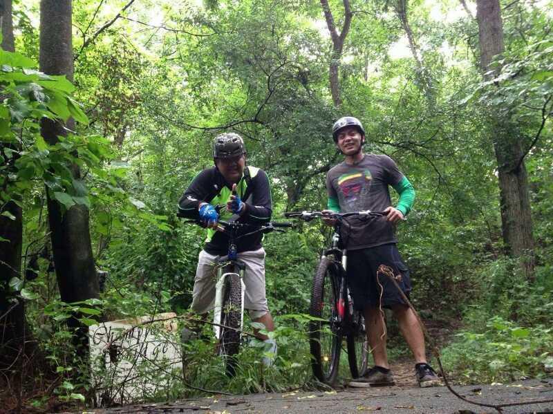 Two mountain bikers posing for a photo in a lush green forest. They are standing beside their bikes, wearing helmets and biking gear. The background is filled with dense foliage and trees, creating a vibrant outdoor atmosphere. Hobby Park mountain bike trail.
