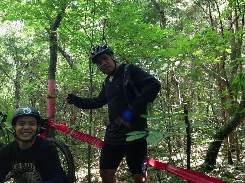 Two mountain bikers posing in a wooded area. One biker is wearing a helmet and a black long-sleeve shirt, while the other is in a black t-shirt and helmet. They are surrounded by green foliage, with a pink ribbon marking a trail in the background. Hobby Park mountain bike trail.