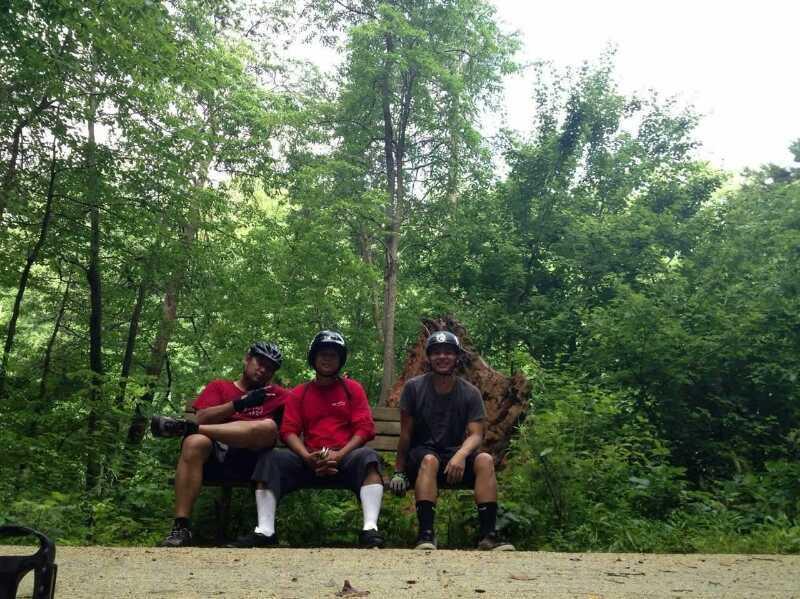 Three individuals wearing helmets and casual clothing are seated on a wooden bench in a lush green forest. The scene captures a moment of relaxation amidst trees and greenery, with sunlight filtering through the leaves. Salem Lake mountain bike trail.