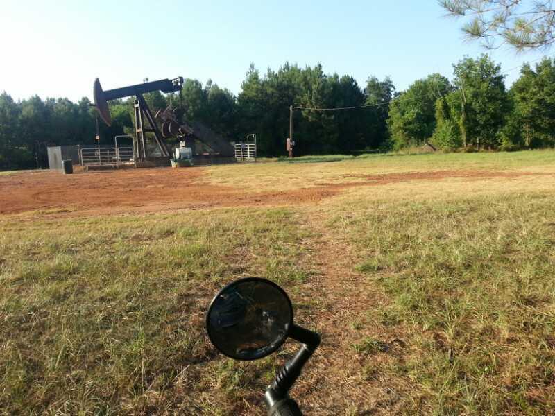 A rural oil field setting with a pumping unit in the background, surrounded by green trees. In the foreground, part of a motorcycle