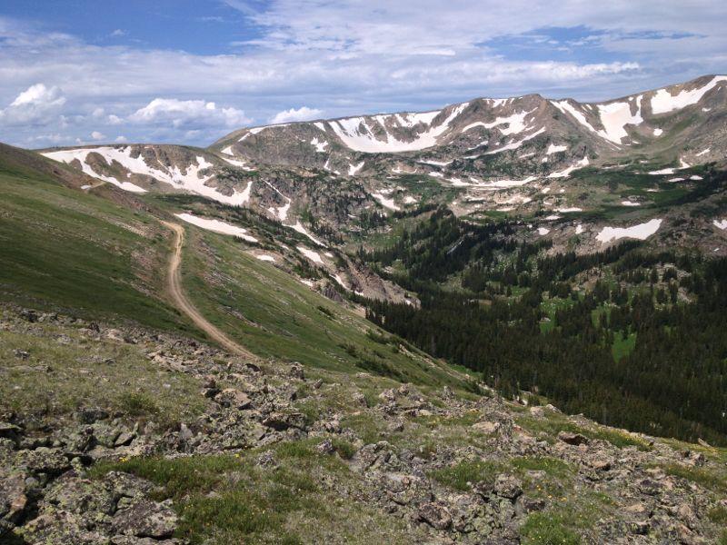 Mountain landscape featuring rolling green hills, rocky terrain, and snow-capped peaks under a partly cloudy sky. A dirt path winds through the scene, leading into the distant valley filled with trees and lush greenery. Rollins Pass mountain bike trail.
