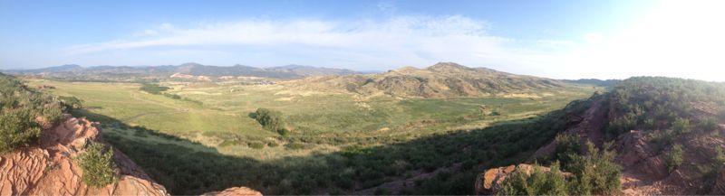 A panoramic view of a scenic landscape featuring rolling hills and green valleys under a clear blue sky. The foreground includes rocky outcrops and shrubs, while distant mountains create a serene backdrop. The image captures the beauty of nature in a wide, open space. Devil's Backbone mountain bike trail.