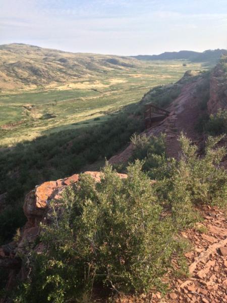 A scenic view of a valley surrounded by rolling hills and cliffs, featuring green vegetation and red rock formations in the foreground. The landscape captures the beauty of nature under a clear sky. Blue Sky mountain bike trail.