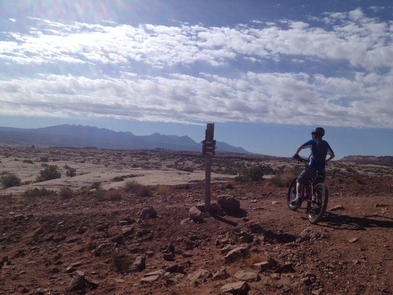 A person on a mountain bike pauses on a rocky trail, gazing at a distant mountain range under a partly cloudy sky. A wooden trail sign stands nearby, indicating directions in the rugged terrain. Moab Brand Trails mountain bike trail.