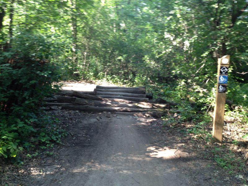 A forest path blocked by several logs, with a trail sign indicating nearby recreational activities. Lush green foliage surrounds the area, providing a natural setting. Lebanon Hills mountain bike trail.