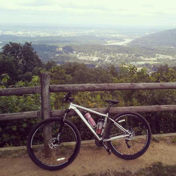 A mountain bike resting against a wooden fence, overlooking a scenic view of valleys and hills in the background, with a city visible in the distance under a partly cloudy sky. A water bottle is attached to the bike's frame. Raccoon Mountain Trail Network mountain bike trail.