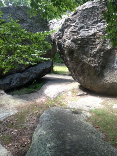 A scenic pathway between two large boulders, surrounded by lush greenery. Sunlight filters through the leaves overhead, illuminating the rocky terrain and creating a tranquil outdoor setting. Lincoln Woods mountain bike trail.