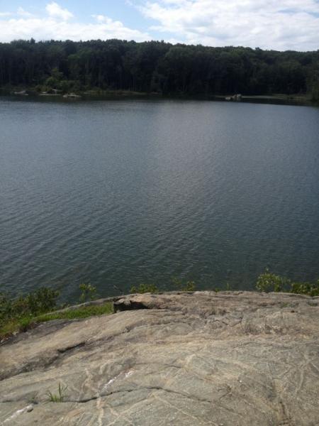 A tranquil view of a lake surrounded by lush greenery, captured from a rocky ledge. The water appears calm with gentle ripples, reflecting the blue sky and scattered clouds above. Lincoln Woods mountain bike trail.