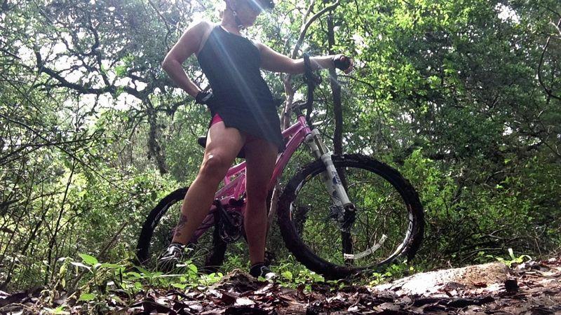 A person in a black tank dress stands confidently beside a pink mountain bike on a dirt path surrounded by greenery. Sunlight filters through the trees, creating a vibrant outdoor scene. Mud Creek mountain bike trail.