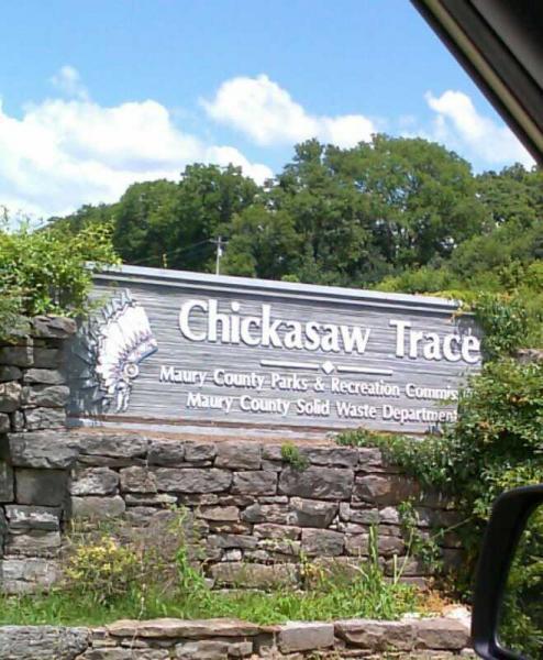 A weathered wooden sign reading "Chickasaw Trace," surrounded by greenery, with additional text indicating it is part of Maury County Parks & Recreation Commission and Maury County Solid Waste Department. The background features a partly cloudy blue sky and trees. Chickasaw Trace mountain bike trail.