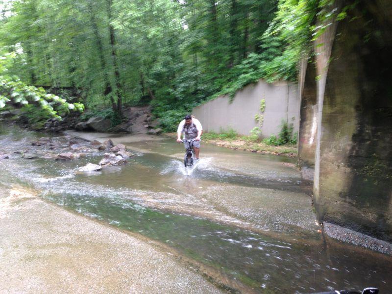 A person riding a bicycle through a shallow stream surrounded by lush greenery and rocky terrain. Water splashes around the bike as they navigate the natural trail. Buttermilk mountain bike trail.