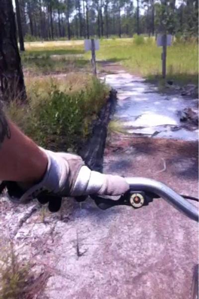 A close-up view of a person's hand wearing a glove holding a bicycle handlebar, with a pathway ahead lined by grass and trees in the background. The road appears slightly worn, and there are signs visible in the distance. Smith Lake mountain bike trail.