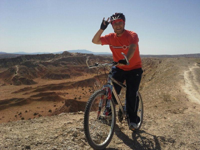 A person in a bright orange shirt and helmet stands next to a mountain bike, smiling and making a hand gesture, with a vast, rocky landscape and blue sky in the background. White Ridge Bike Trails mountain bike trail.