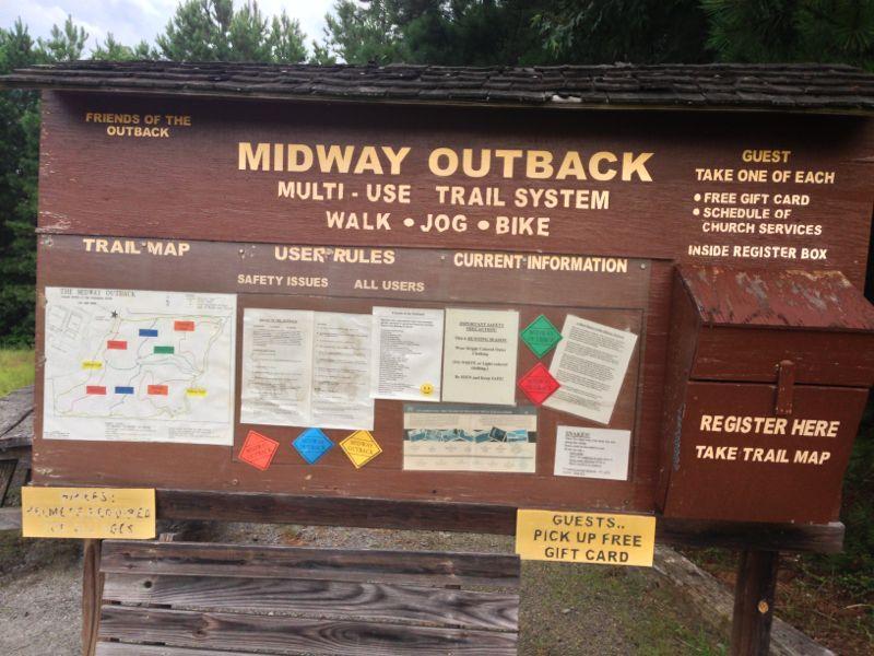 Signboard for Midway Outback, displaying information about the multi-use trail system for walking, jogging, and biking. Includes a trail map, user rules, safety issues, and announcements about free gift cards and church service schedules. A register box for guests is visible on the right side. Midway Outback mountain bike trail.