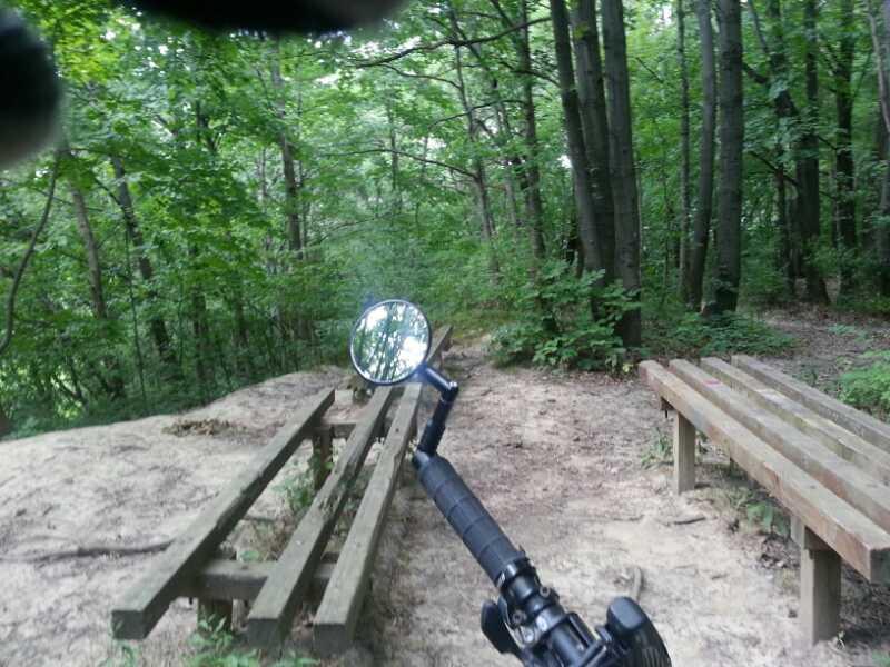 A view of a forested path, taken from a person's perspective holding a handlebar with a mirror. There are wooden benches on either side of the path, and dense greenery surrounds the area, with tall trees providing a natural canopy. Murdock Park Trail mountain bike trail.