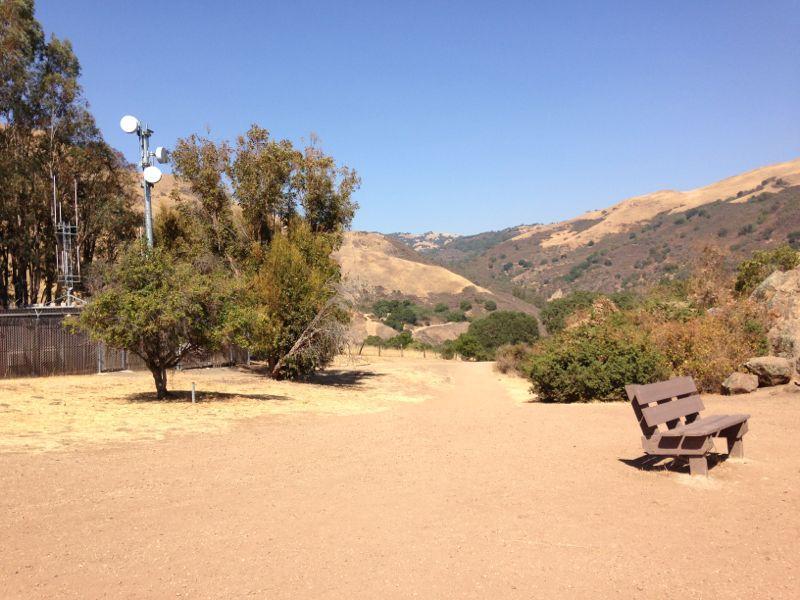 A dusty pathway lined with trees, leading into a serene landscape of rolling hills under a clear blue sky. On the right, a wooden bench is positioned along the path, inviting visitors to rest and enjoy the surrounding natural beauty. In the background, a telecommunications tower is partially visible among the trees. Alum Rock County Park mountain bike trail.