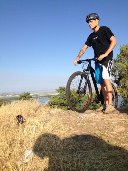 A young man in a black helmet and a black t-shirt stands on a rocky ledge with a mountain bike. He is positioned on the edge of a natural area with tall grass and trees, overlooking a river and blue sky in the background. The sunlight casts a shadow on the ground beside him. Glendo State Park mountain bike trail.