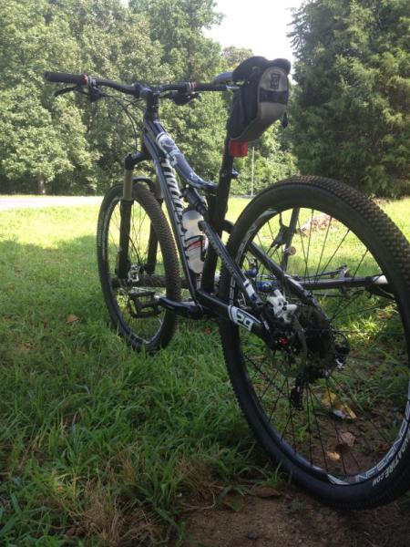 A close-up view of a mountain bike parked on grassy ground, with a blurred background of trees and a road. The bike features a sleek black frame, a water bottle holder, and a small bag attached to the handlebars. The tires are rugged, suitable for off-road cycling. Hobby Park mountain bike trail.