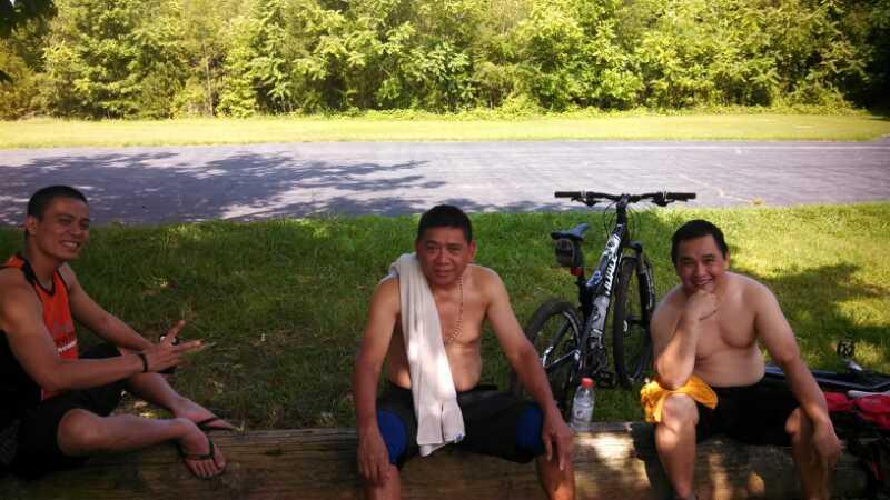 Three men sitting on a log by a road, enjoying a break. They are shirtless and wearing shorts, with one man wearing a towel around his neck. A bicycle is parked nearby, and the background features greenery and trees. Hobby Park mountain bike trail.