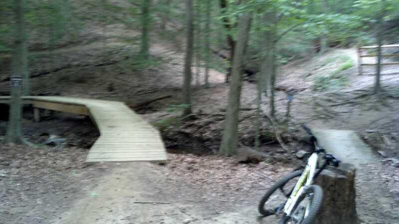 A blurred image of a wooded area featuring a wooden bridge over a small creek. To the right, a bike is propped against a tree stump. The scene includes dense vegetation and a trail that leads through the forest. Fountainhead Regional Park mountain bike trail.