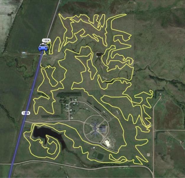 Aerial view of a regional area with a winding route highlighted in yellow, illustrating a driving path that navigates through open land and a small community, with a blue car icon indicating the starting point near a highway. Adventist Academy Trail mountain bike trail.