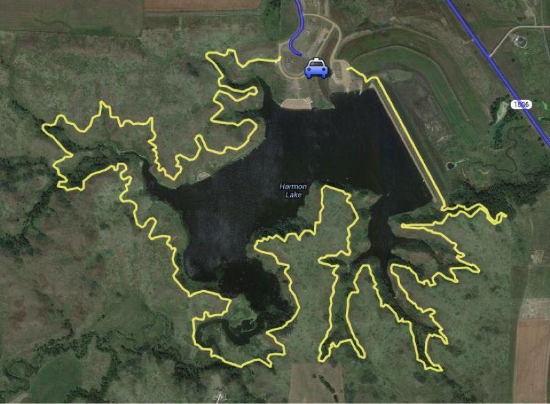 A satellite image showing Harmon Lake, outlined in yellow, with a nearby parking area marked by a blue car icon. The landscape includes greenery and agricultural fields surrounding the water body. Harmon mountain bike trail.