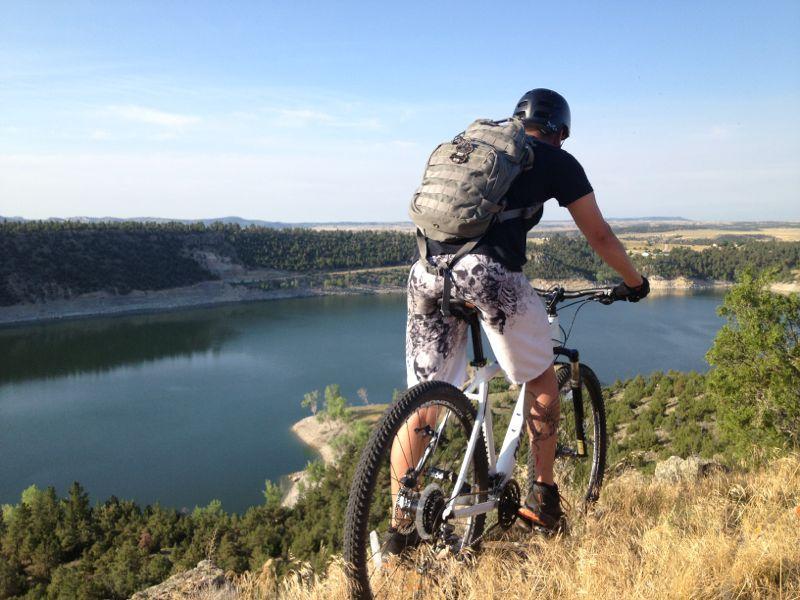 A person wearing a helmet and a backpack is standing on a mountain bike, overlooking a tranquil lake surrounded by greenery and hills. The scene is set under a clear blue sky on a sunny day. Glendo State Park mountain bike trail.