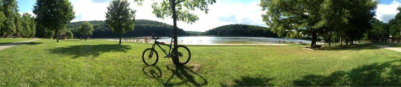 Panoramic view of a serene lakeside park, featuring a bicycle leaning against a tree on lush green grass. The lake reflects the blue sky and surrounding hills, with people enjoying the water in the distance. Greenbrier State Park mountain bike trail.