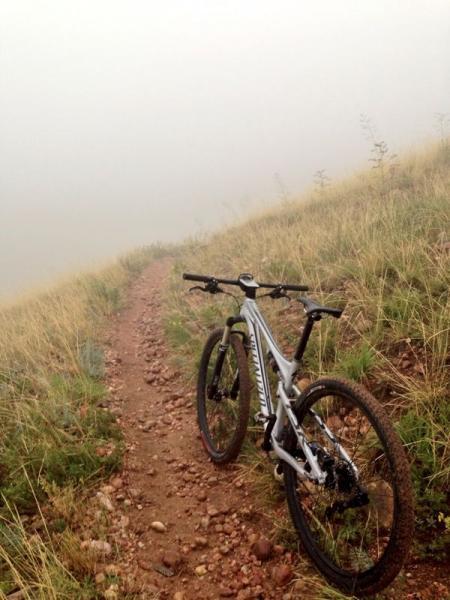 A mountain bike resting on a rocky trail, surrounded by tall grass and fog, creating a serene and mysterious atmosphere. The visibility is limited due to the fog, emphasizing the remote natural setting of the trail. Green Mountain mountain bike trail.