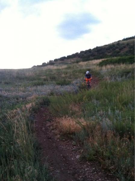 A child in orange riding a bicycle along a dirt trail through a grassy field, with a hilly landscape in the background under a cloudy sky. Green Mountain mountain bike trail.