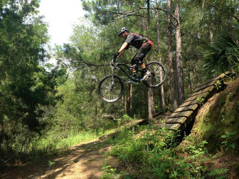 A mountain biker mid-air while jumping off a wooden ramp in a forested area, surrounded by trees and greenery. The rider is wearing a helmet and protective gear, showcasing a dynamic action pose against a natural backdrop. Santos mountain bike trail.
