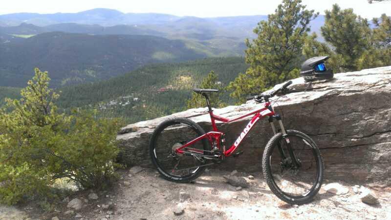 A red mountain bike parked on a rocky outcrop with a black helmet resting on the rocks. The backdrop features a scenic view of green mountains and valleys under a blue sky. Pines frame the sides of the image, adding to the outdoor adventure atmosphere. Elk Meadow mountain bike trail.