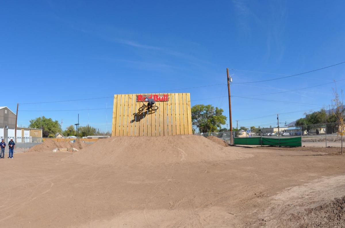 A cyclist performing a trick on a vertical wooden ramp, branded with the Red Bull logo, set against a clear blue sky. In the foreground, two spectators watch from a dirt surface. The background features buildings and utility poles, typical of an outdoor biking venue. North Valley Bike Park mountain bike trail.