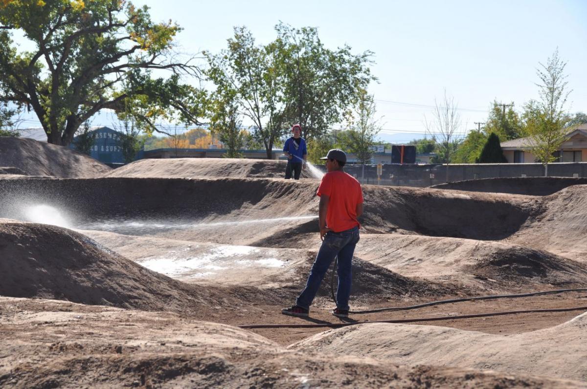 Two individuals are working on a dirt track; one is spraying water onto the terrain while the other observes. The scene features a landscape of dirt mounds under a clear blue sky, with trees and buildings in the background. North Valley Bike Park mountain bike trail.