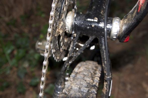 Ragley Blue Pig: Close-up view of a bicycle's drivetrain, featuring a muddy chain, rear cog, and wheel. The background shows blurred greenery, indicating an off-road environment.