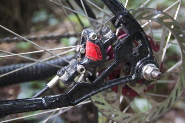 Ragley Blue Pig: Close-up view of a bicycle's rear brake assembly, featuring a black frame with a red brake caliper, metallic components, and a visible disc brake rotor, set against a blurred background of greenery.