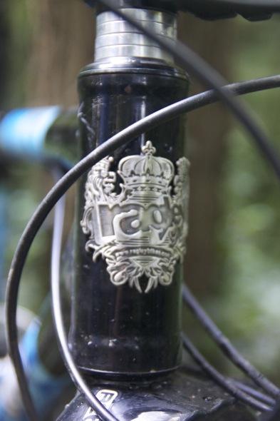 Ragley Blue Pig: A close-up shot of a bike's head tube featuring a decorative silver logo with a crown, partially obscured by black bike cables. The background shows a blurred green forest setting.