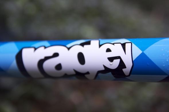 Ragley Blue Pig: Close-up of a blue bicycle frame featuring the logo "Radley" in bold, black lettering on a patterned background.