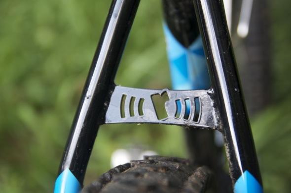 Ragley Blue Pig: Close-up view of a bicycle frame's rear triangle, featuring a metallic bridge with ventilation slots and blue accents. The background is blurred, showcasing green grass.