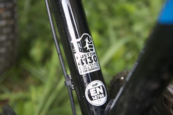 Ragley Blue Pig: A close-up image of a bicycle frame's downtube, featuring a black finish. The frame displays various decals, including "CUSTOM 4130" and "Heat Treated," alongside a certification label marked "EN 14766." The background shows blurred greenery, indicating an outdoor setting.