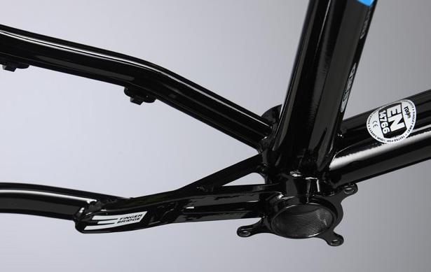 Ragley Blue Pig: Close-up of a black bicycle frame's bottom bracket area, highlighting the tubing and fastening points. The image features a glossy finish and includes certification stickers visible on the frame. The background is a neutral gray, emphasizing the frame's details.