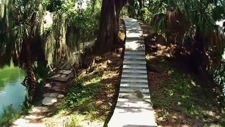 A winding stone pathway leads through a lush green area, bordered by trees and vegetation, next to a body of water. The pathway ascends gently, inviting visitors to explore the natural surroundings. Loyce E. Harpe Park mountain bike trail.