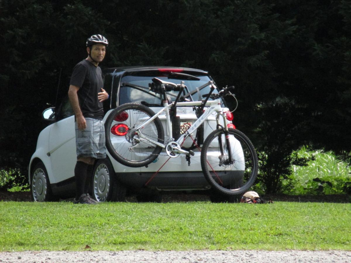 Orbea Alma 29 World Cup: A man wearing a black helmet, black shirt, and plaid shorts stands next to a small white car with a mountain bike attached to the back. The scene is set in a grassy area with trees in the background, suggesting an outdoor setting.