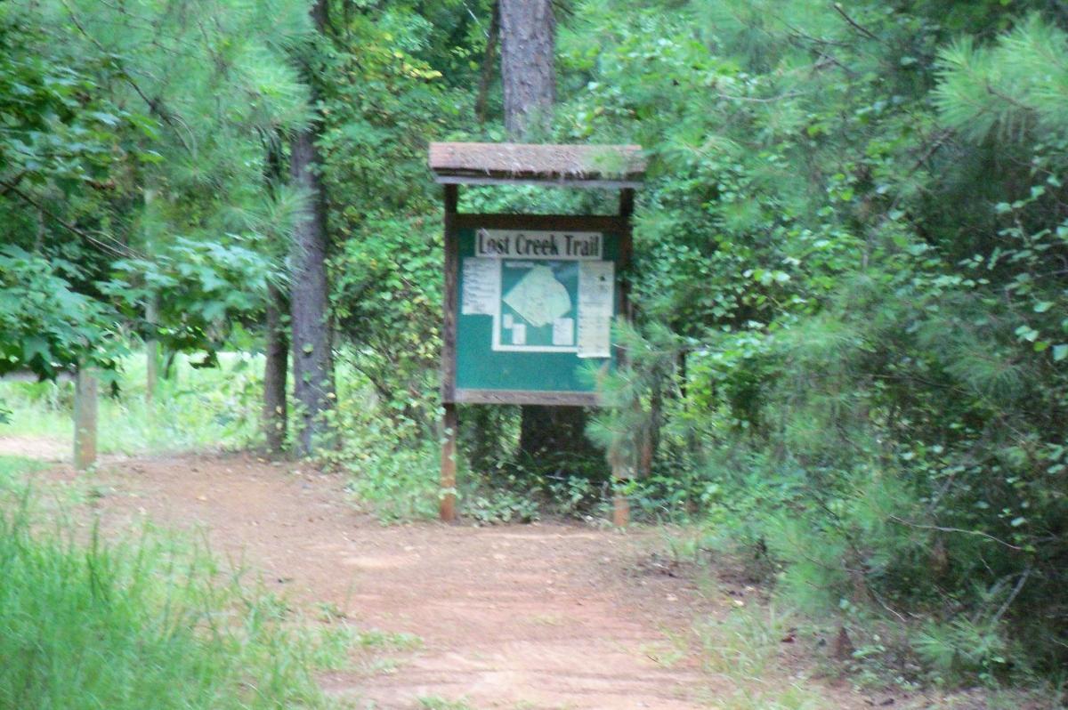 A wooden sign marking the entrance to the Lost Creek Trail, surrounded by lush greenery and trees. The sign includes a trail map and information about the area. A dirt path leads into the forest. Harbison State Forest mountain bike trail.