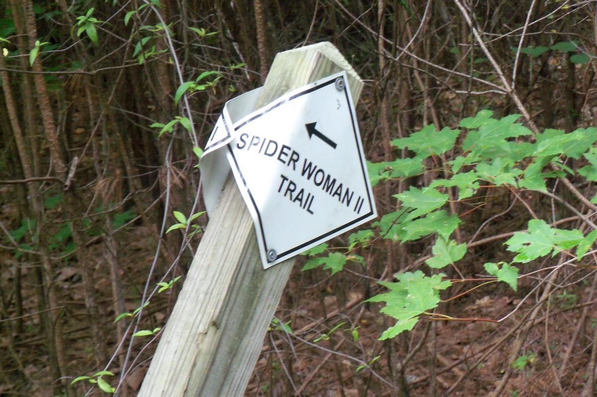 A wooden post with a directional sign indicating the "Spider Woman II Trail," surrounded by greenery and trees in a forested area. Harbison State Forest mountain bike trail.